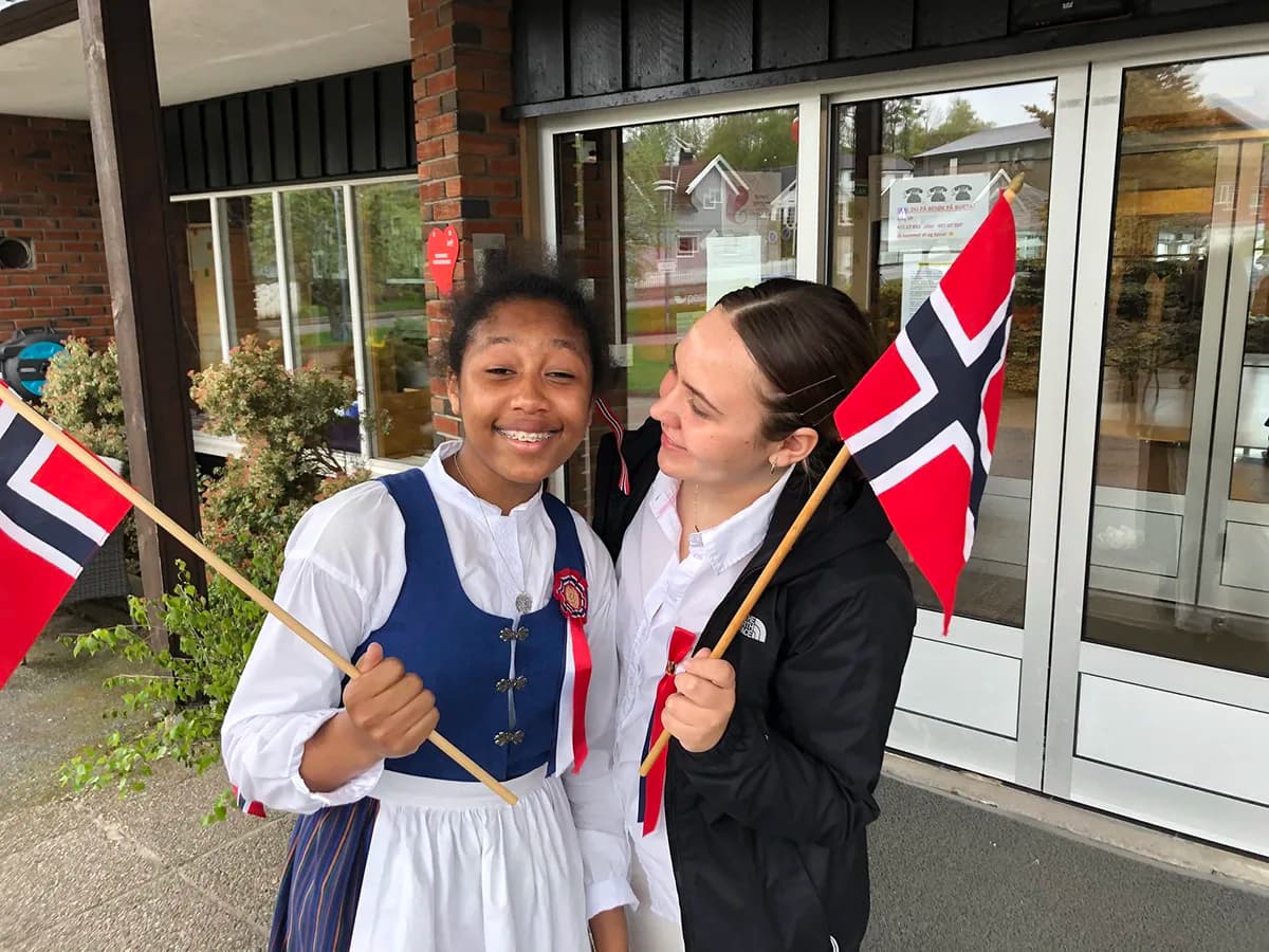 Due ragazze sorridono insieme con le bandiere norvegesi, una in abito tradizionale norvegese, vivendo un’esperienza di scambio culturale durante l’anno all’estero in Norvegia.